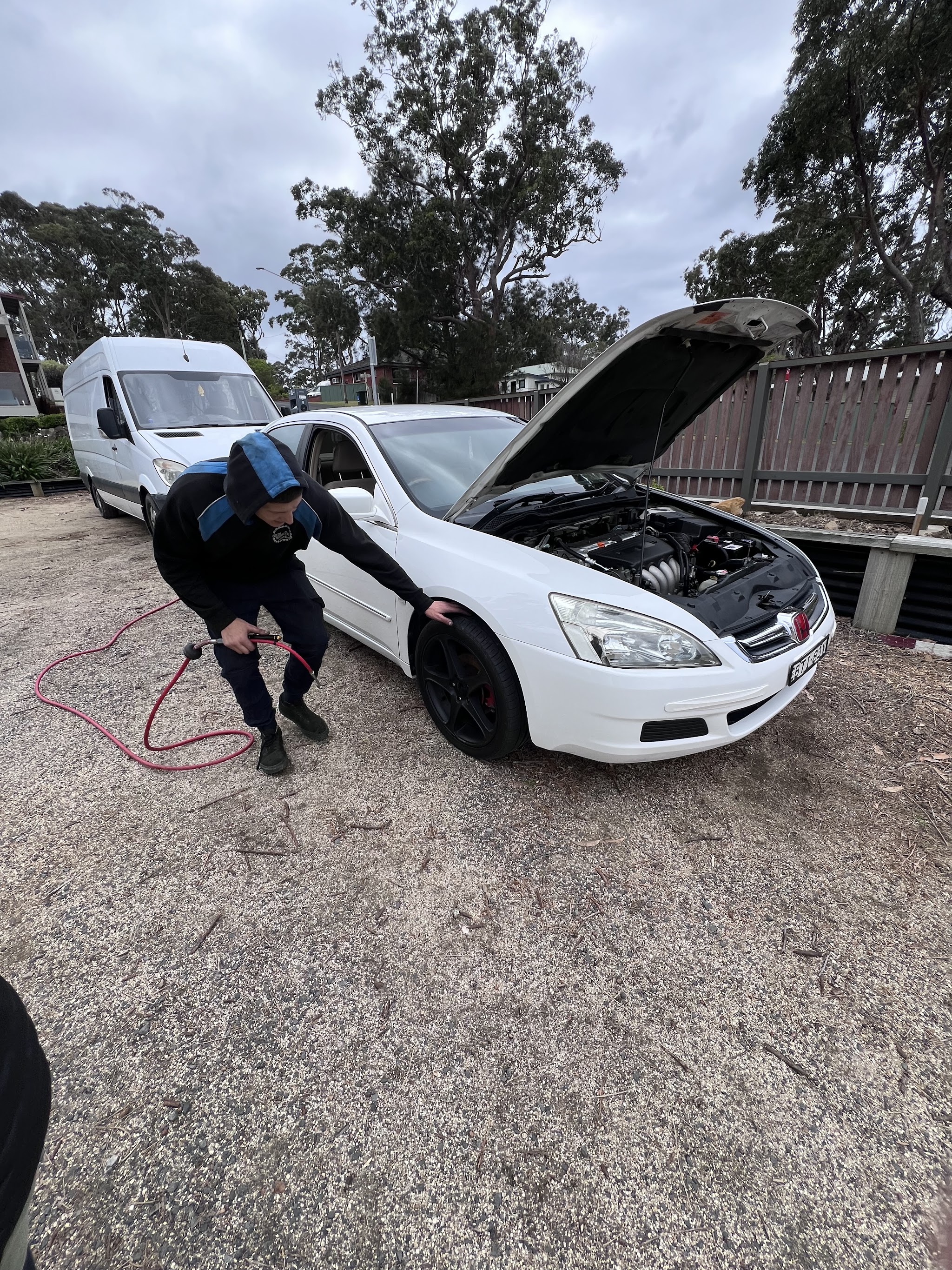A mobile mechanic checking tyre pressure on a customer's white car, bonnet up, with the Endless Automotive van behind