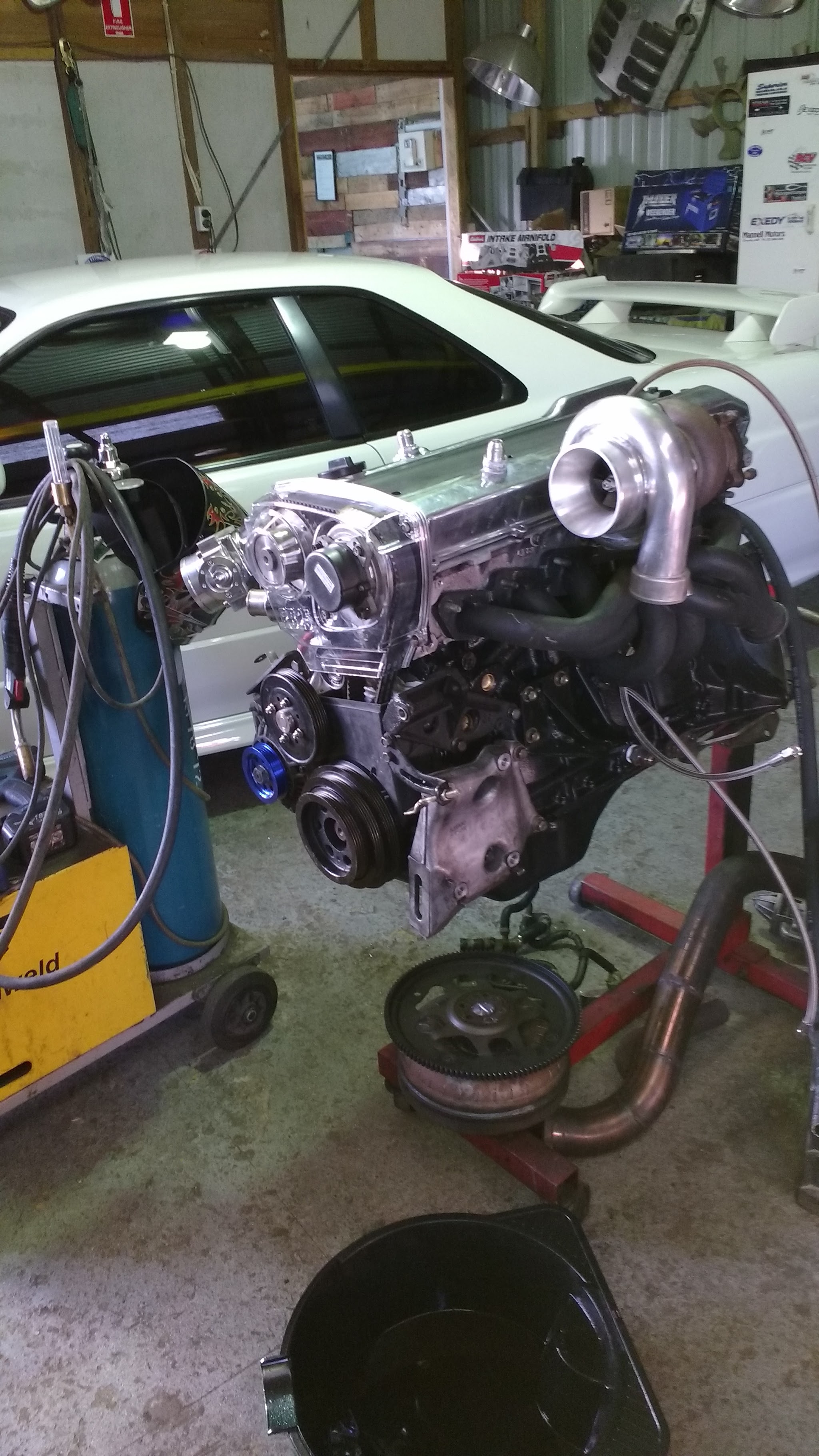 A built turbo engine on an engine stand in the workshop, a white sports car in the bay behind